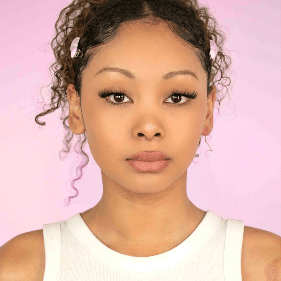 Front-facing portrait of a woman wearing long, flared, pointed cat-eye false eyelashes against a light pink background.
